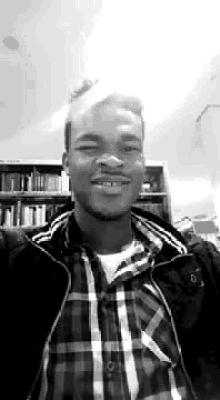 a black and white photo of a man in a plaid shirt and jacket smiling in front of a bookshelf .