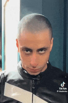 a young man with a shaved head is wearing a leather jacket and making a funny face .