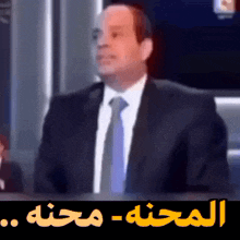 a man in a suit and tie is sitting in front of a screen with arabic writing on it