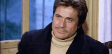 a man with a mustache is sitting in front of a window wearing a turtleneck sweater and a coat .