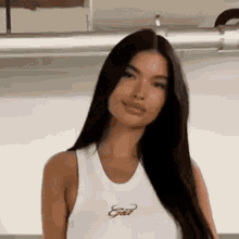 a woman with long black hair is wearing a white tank top and smiling at the camera .