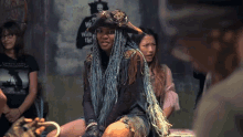 a woman with blue braids and a pirate hat is sitting in front of a wall that says we 're with the