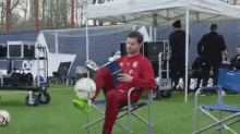 a man is sitting in a chair on a soccer field reading a book while holding a soccer ball .