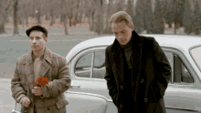 two men are standing next to a car one is holding a red flower