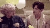 two young men are sitting at a table with balloons in the background