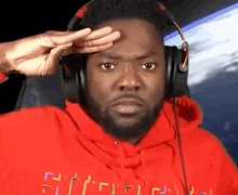 a man wearing headphones and a red hoodie salutes while looking at the camera .