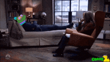 a woman sits in a chair next to a man laying on a couch with nbc written on the bottom of the screen
