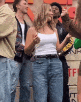 a woman in a white tank top and blue jeans stands in a crowd of people