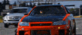 a red and black car is driving down a street next to a white truck .