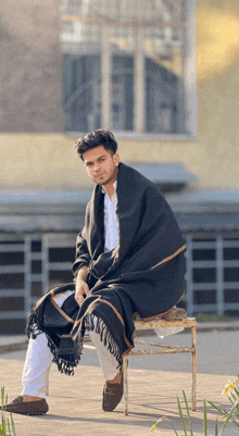 a man wrapped in a black blanket sits on a chair in front of a building