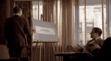 a man in a suit is giving a presentation to another man in an office while holding a sign that says admiral .