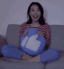 a woman is sitting on a couch holding a blue pillow with a thumbs up symbol on it