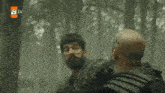 a man with a beard is holding another man 's arm in a forest .