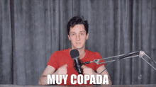 a man in a red shirt stands in front of a microphone with the words muy copada above him