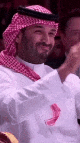 a man with a beard is wearing a keffiyeh and smiling while clapping his hands .