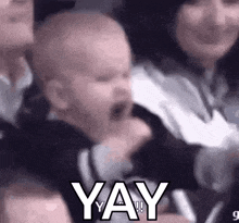 a baby is sitting in a crowd of people with his mouth open and the words `` yay '' written on it .