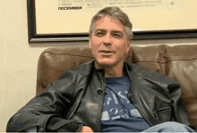a man in a leather jacket is sitting on a couch in front of a framed picture that says december