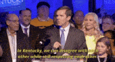 a man in a suit and tie stands at a podium with a crowd behind him and says in kentucky