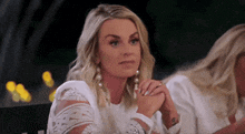 a woman in a white dress is sitting at a table with her hands folded and looking at the camera .