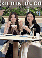 two women are sitting at a table in front of a sign that says ' alain duca ' on it
