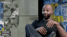 a bald man with a beard is sitting in a chair with his legs crossed and talking to someone .