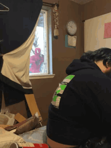 a man in a black hoodie with the letter s on the back sits in a messy room