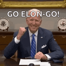a man in a suit and tie is sitting at a desk with a fist in the air and says go elon-go .