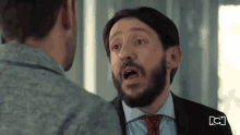 a man in a suit and tie is talking to another man in a grey shirt .