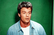 a man wearing a denim shirt and white shirt stands in front of a green wall