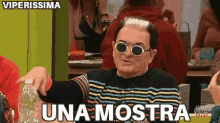 a man wearing sunglasses and a striped shirt is sitting at a table and pointing at the camera .