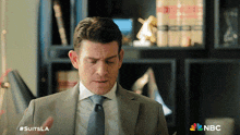a man in a suit and tie is standing in front of a bookshelf with the nbc logo on the bottom