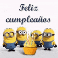 a group of minions standing around a cupcake with candles on it and the words feliz cumpleanos written above them .
