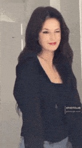 a woman wearing a black top and blue jeans is standing in front of a white wall .