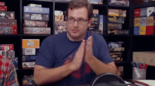 a man wearing glasses is sitting at a table with his hands folded in front of a shelf full of board games .