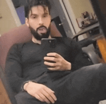 a man with a beard is taking a selfie with his phone while sitting in a chair