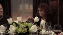 a woman with curly hair sits at a table with flowers and glasses of wine