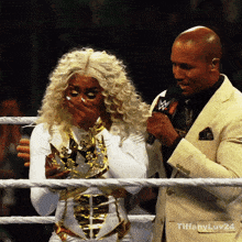 a man in a suit stands next to a woman in a wrestling ring with tiffany.lv24 written on the bottom right