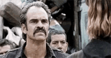 a man with a mustache is talking to a woman in front of a group of people .
