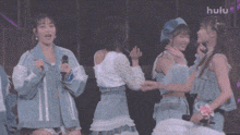 a group of girls are standing on a stage holding hands and singing into microphones while hulu is displayed in the background .