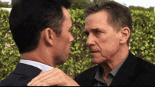 two men in suits are looking at each other in front of a bush