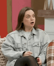 a woman in a denim jacket is sitting on a couch and making a surprised face .
