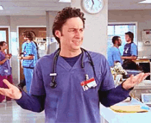 a man wearing a blue scrub top with a stethoscope around his neck stands in a hospital room