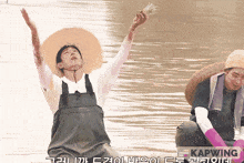 a man wearing overalls and a hat is standing in the water with his arms in the air and the word kapwing below him