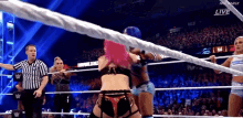 a woman with pink hair is wrestling another woman in a wrestling ring while a referee looks on .