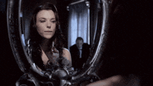 a woman is looking at herself in a mirror with a man in the background