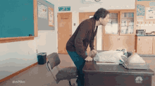 a man is standing in front of a desk in a classroom with a bag on it .