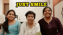 three women are smiling in front of a sign that reads just smile