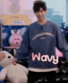 a man wearing a blue sweater is holding a stuffed animal and says wavy .
