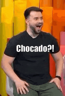 a man wearing a black t-shirt that says chocado is sitting in a chair with his mouth open .
