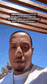 a man wearing a white shirt and a gold chain is standing under a wooden pergola